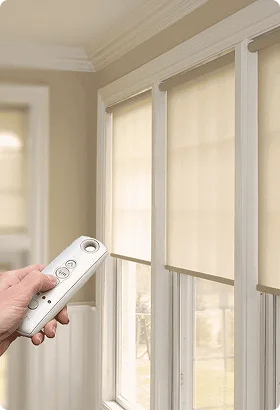 A hand holding a remote control operates motorized beige window shades